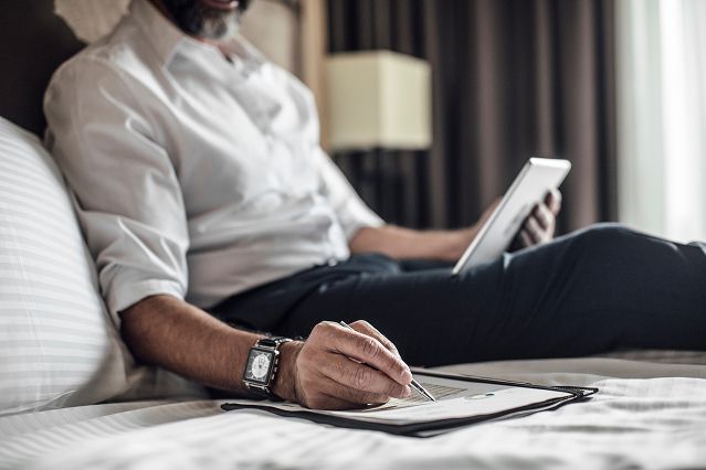 A man comfortably sitting on a bed, utilizing a tablet and pen for writing